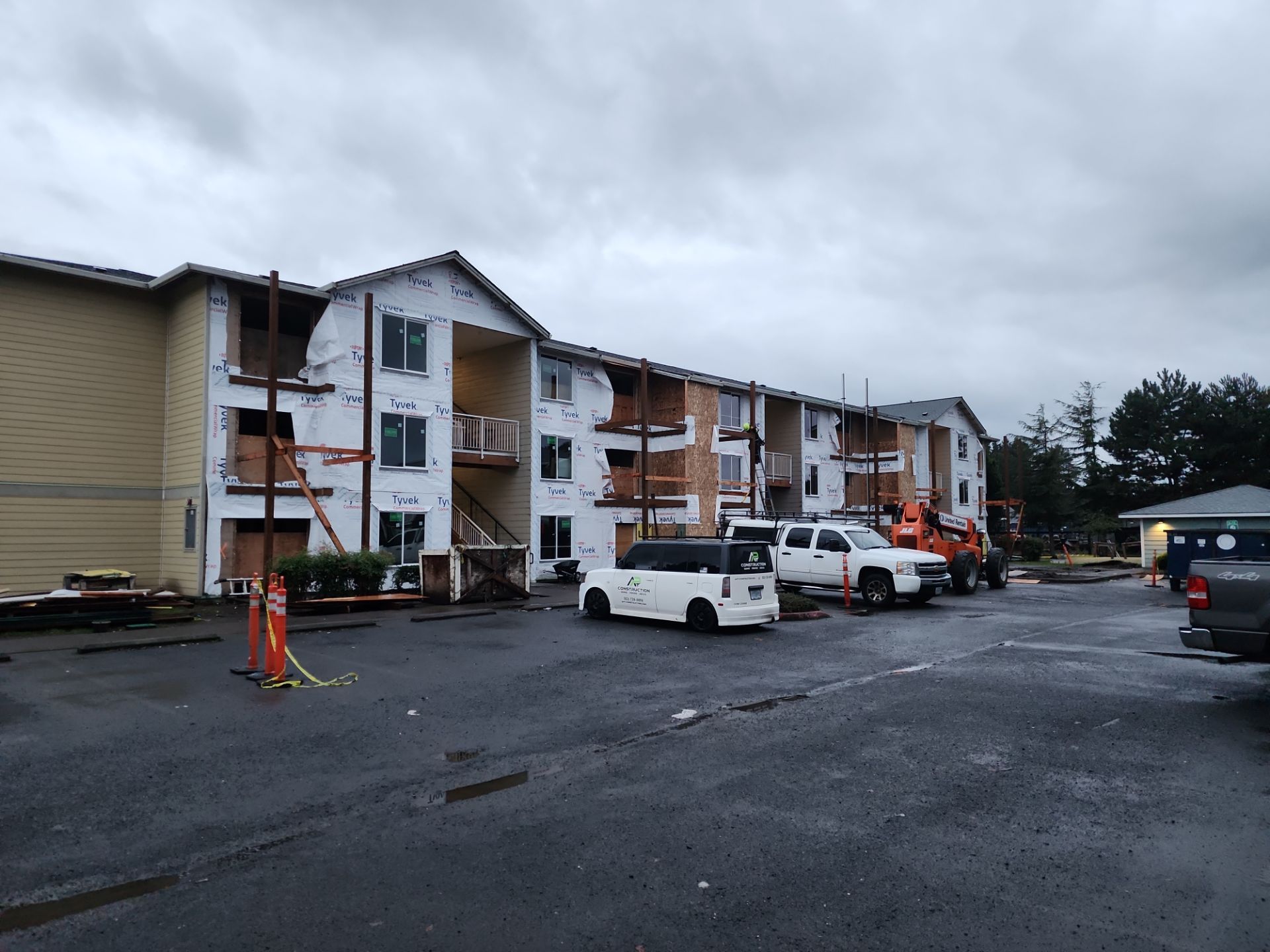 A three-story apartment building under renovation, with exposed framing and construction materials. Vehicles and construction equipment are parked in front on a cloudy day.