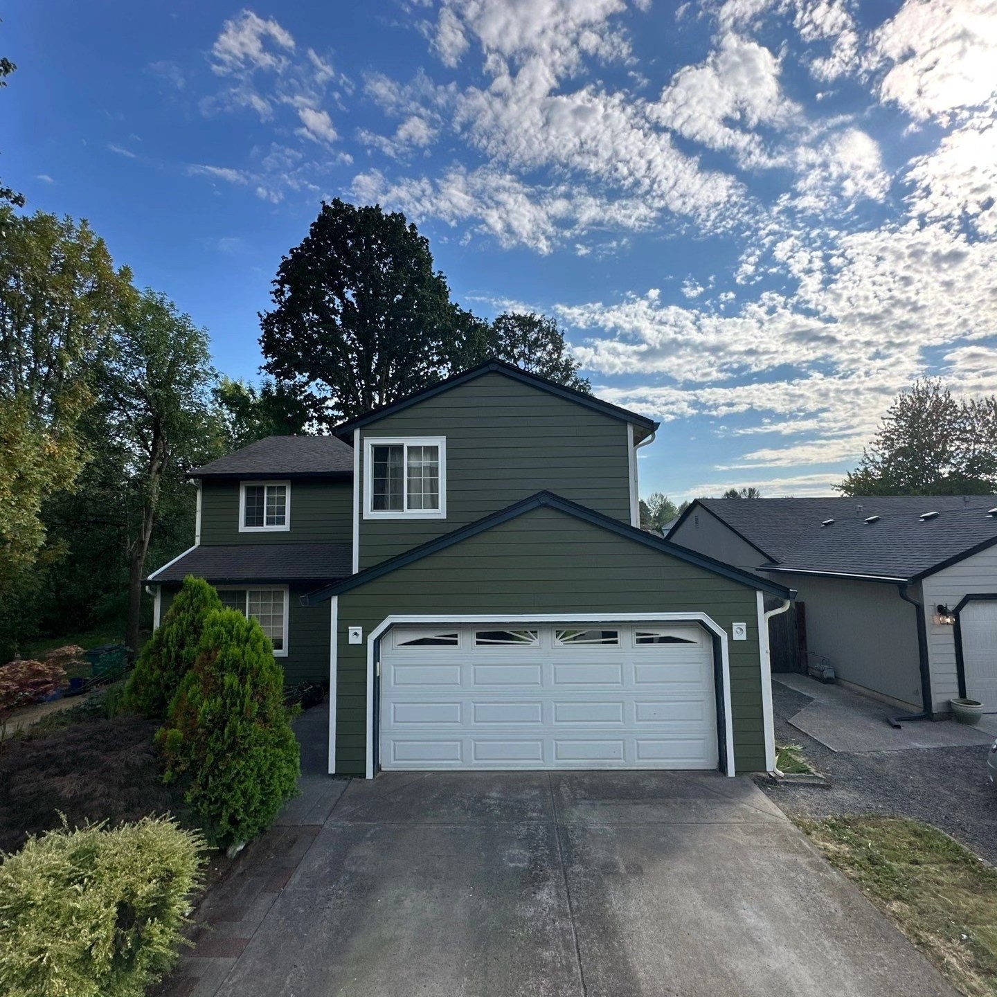 A two-story green house with a white double garage door, a concrete driveway, and surrounding trees under a partly cloudy sky.