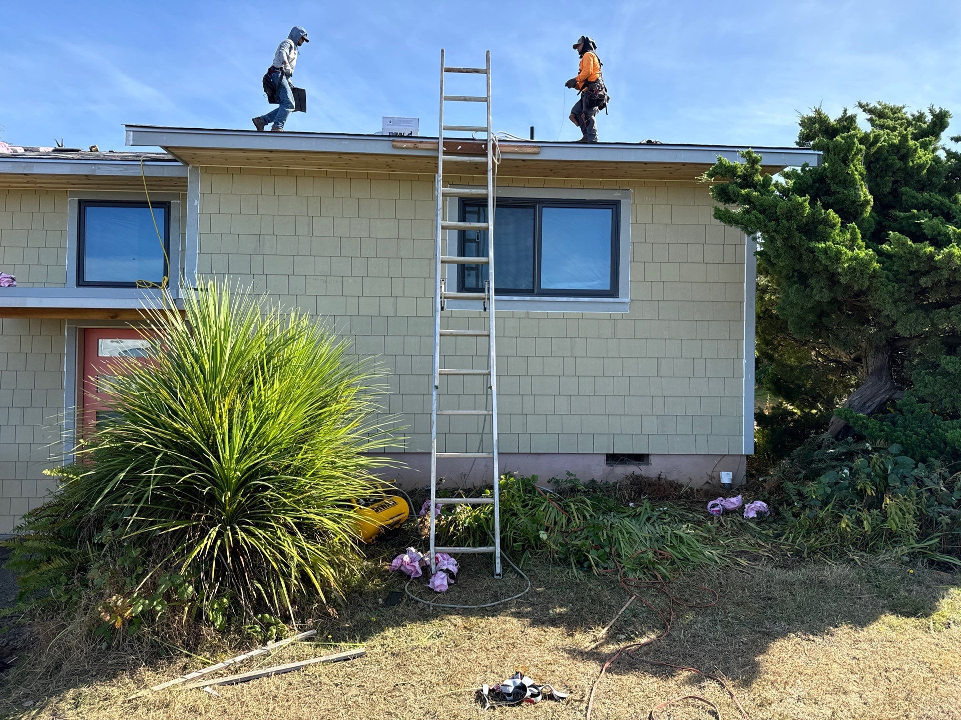 Two workers stand on a house roof near a ladder, with landscaping tools, plants, and flowers visible in the yard below.