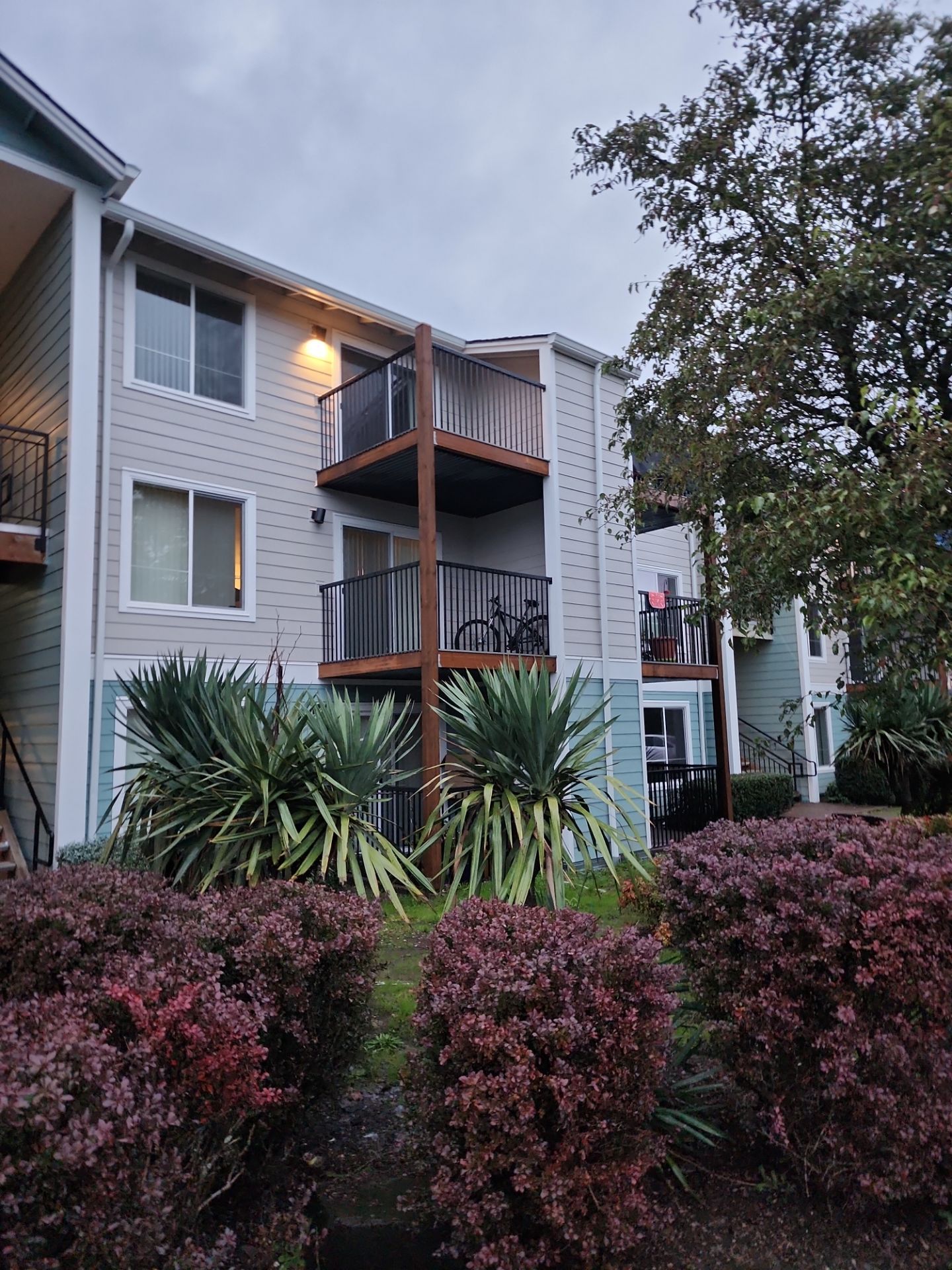 A two-story apartment building with balconies, surrounded by shrubs and trees, under a cloudy sky.