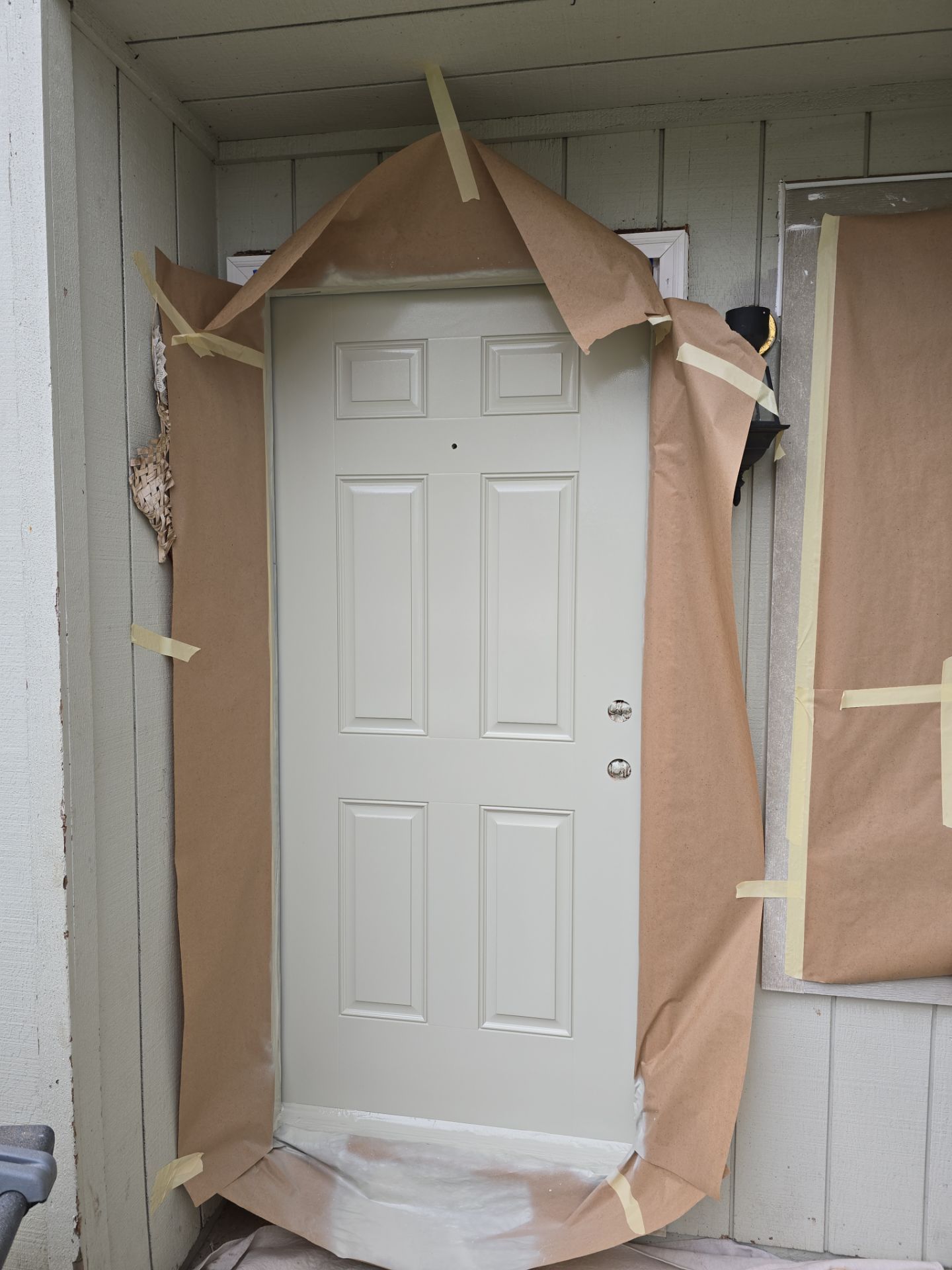 A front door surrounded by brown paper and masking tape, with nearby wall and trim also partially covered, likely prepared for painting or spraying.