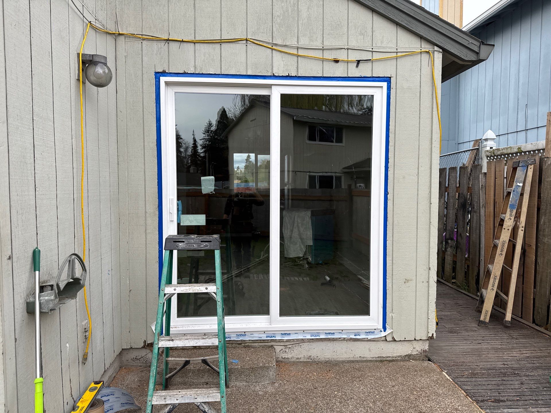 A newly completed door installation features a sliding glass door on a house exterior, with blue tape around the frame and a green step ladder in front. Tools and a garden hose are visible nearby.