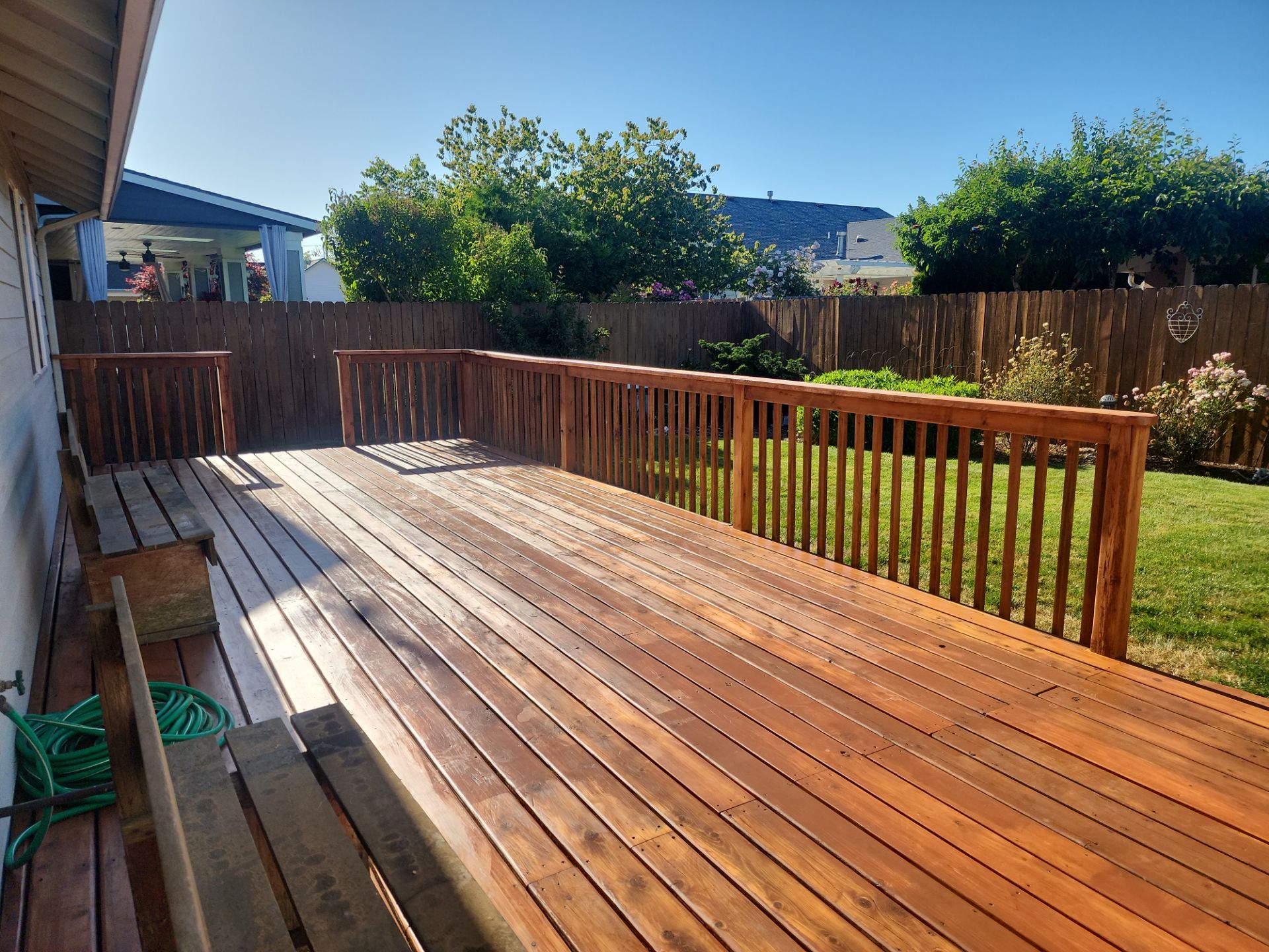 A freshly stained wooden deck extends from a house, bordered by a wooden railing, overlooking a fenced backyard with green grass and shrubs.