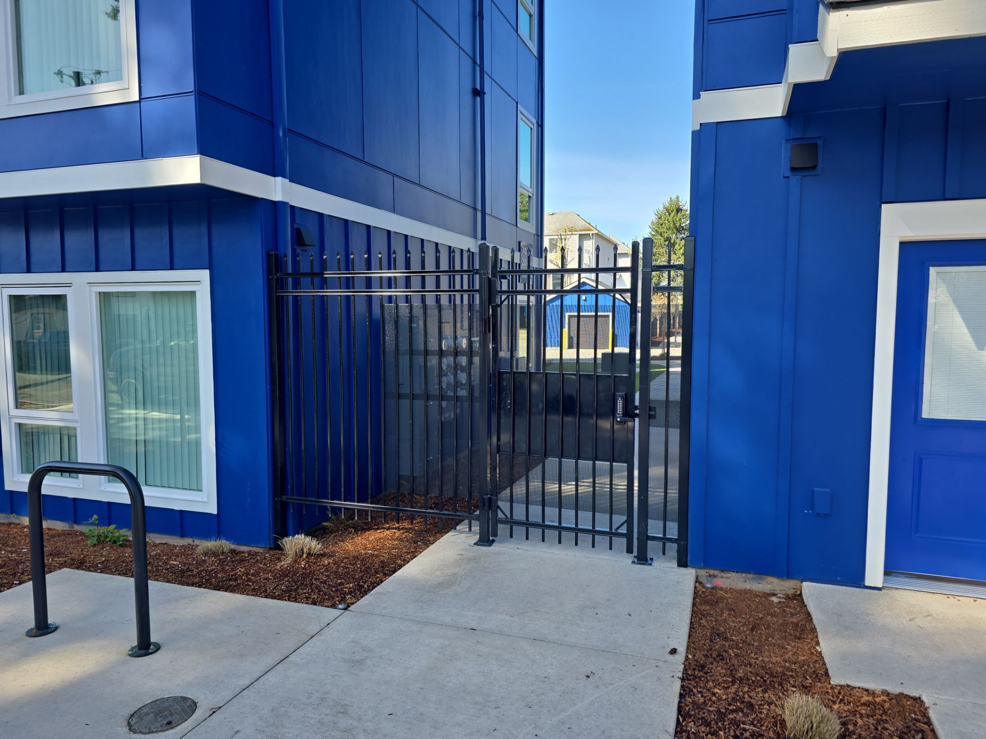 A black metal gate is set between two blue buildings, with a sidewalk and bike rack in the foreground and a small patch of mulch with dry grass.