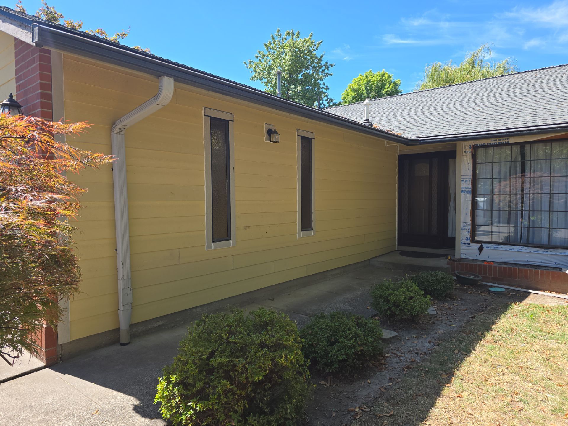 A yellow house exterior with three narrow vertical windows, a dark front door, small shrubs along the walkway, and a clear blue sky.
