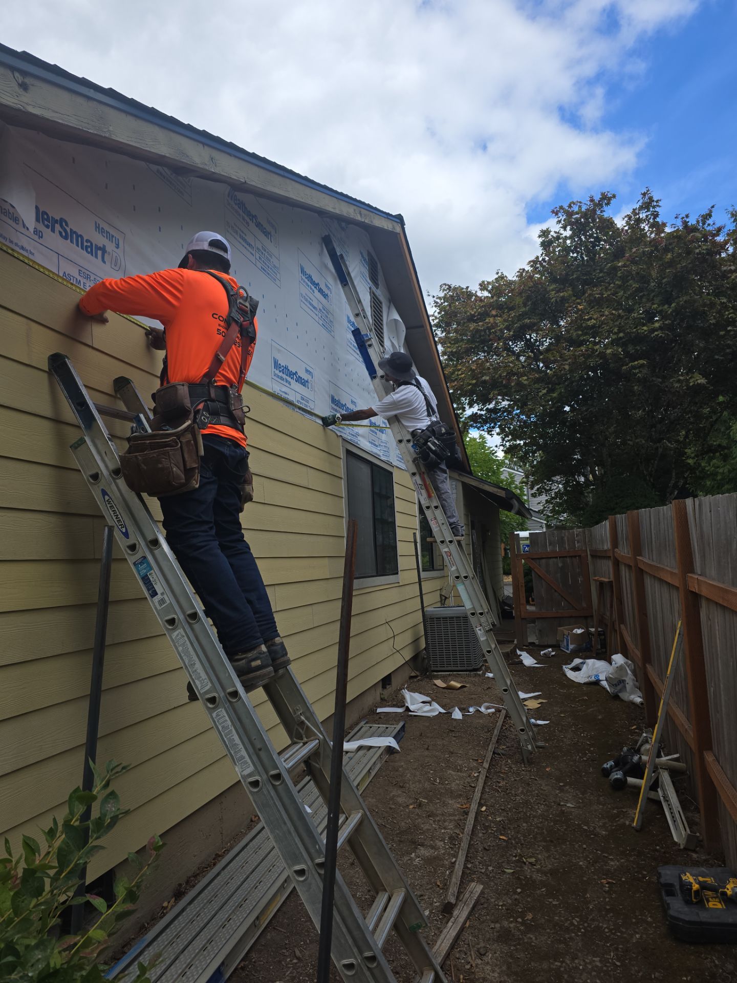 Two construction workers on ladders install siding on a house. Tools and materials are scattered on the ground nearby. The sky is partly cloudy.