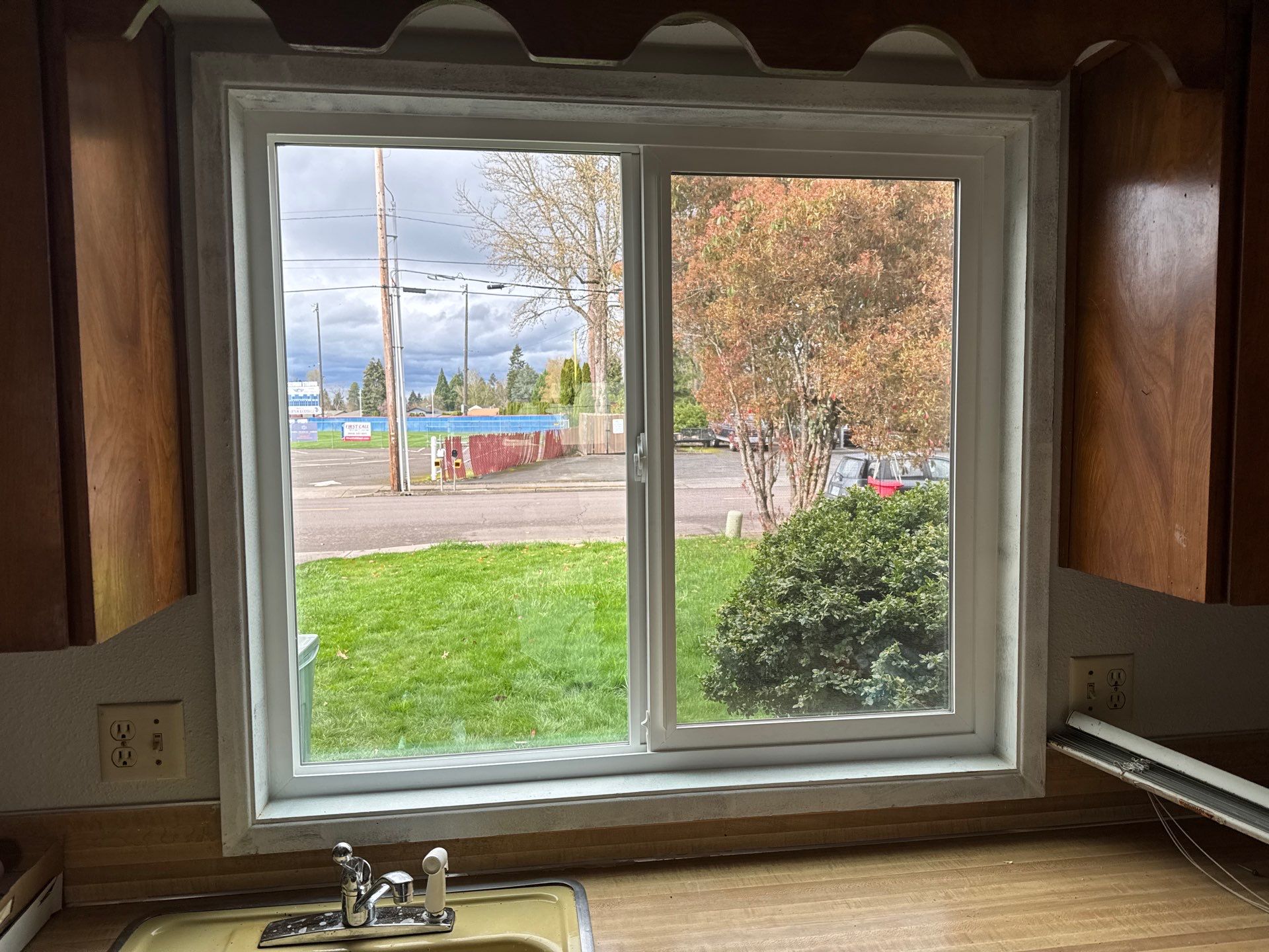 A kitchen window above a sink looks out onto a yard, street, and trees under a cloudy sky. There are power outlets on both sides of the window.