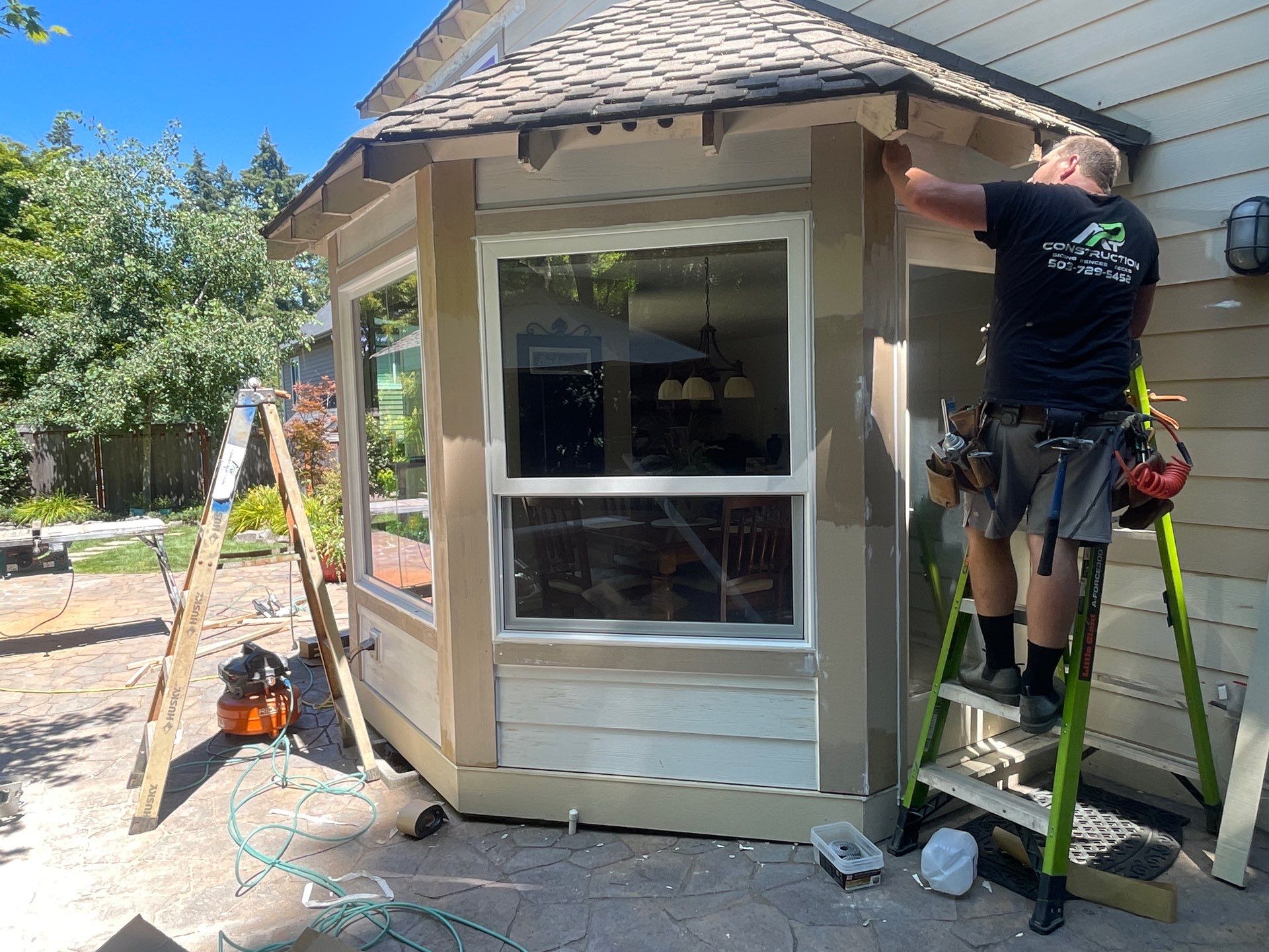 A man stands on a ladder painting the trim of a house's bay window exterior. Tools and equipment are scattered nearby on the patio.